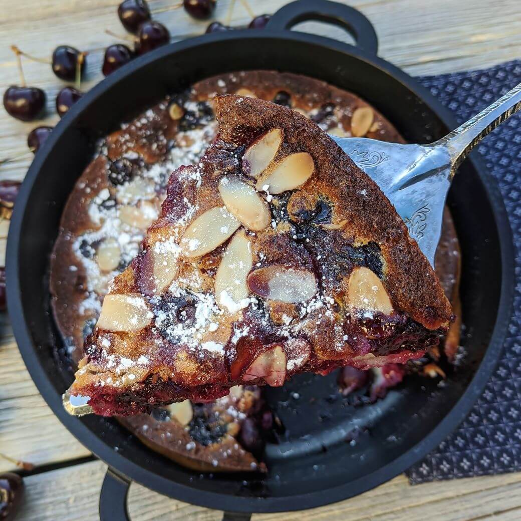 A silver cake server holds a piece of cherry clafoutis topped with powdered sugar and almonds above a pan of the same clafoutis.