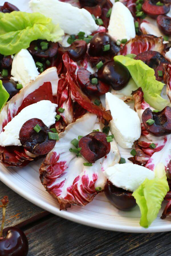 A beautiful salad of grilled radicchio, bright red cherries, green lettuce, and white goat cheese quenelles on a white plate.