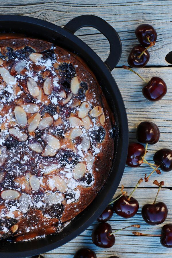 A cast iron pan containing baked cherry clafoutis topped with almonds and powdered sugar surrounded by fresh cherries.