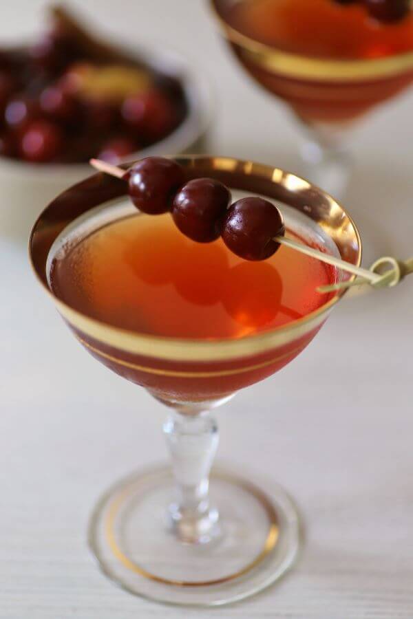 A Manhattan cocktail in a gold rimmed coupe glass garnished with three cherries on a stick.