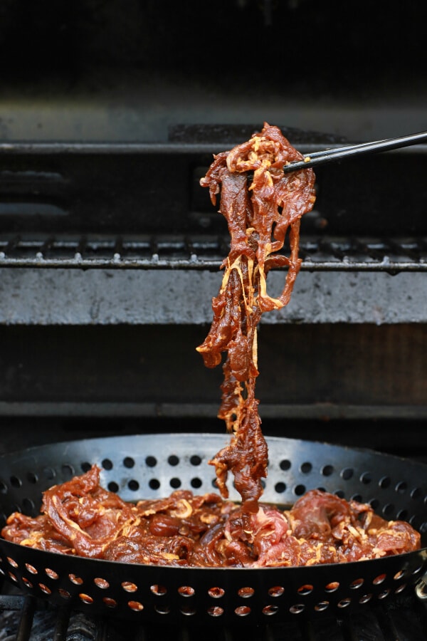 A pair of chopsticks hold marinated raw beef above a grilling basket.