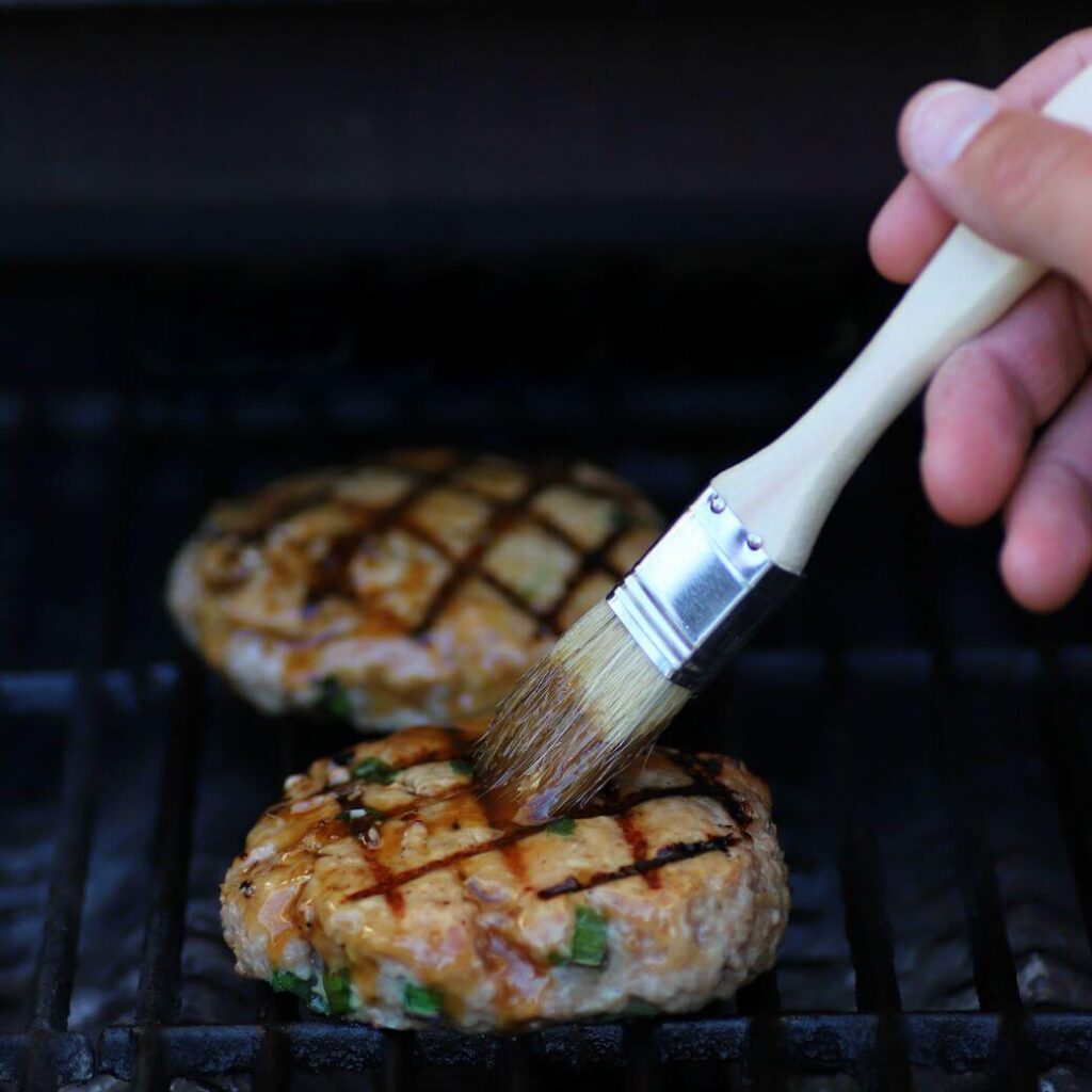 A hand holding a brush while spreading Honey Teriyaki Sauce on Burgers while cooking on a barbecue.