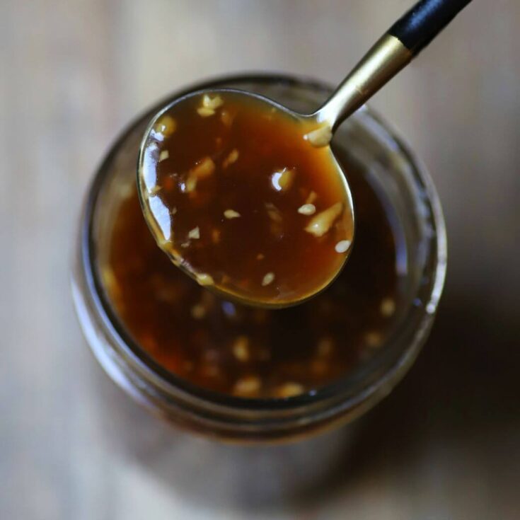 A spoon holds brown Honey Teriyaki Sauce containing chunks of garlic and ginger over a jar of the same sauce.