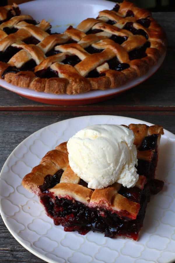 A slice of cherry pie with golden baked lattice top topped with a scoop of vanilla ice cream.