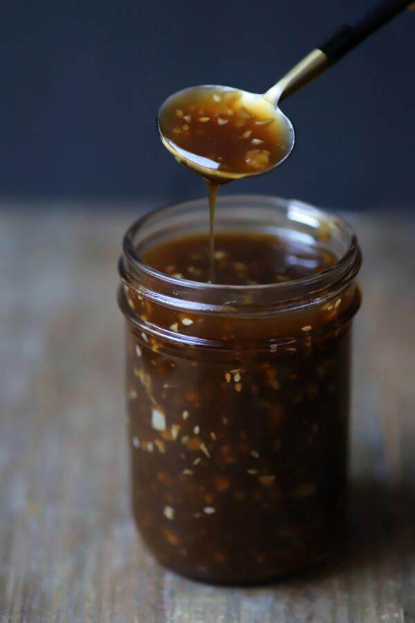 A spoon holds brown Honey Teriyaki Sauce containing chunks of garlic and ginger over a jar of the same sauce.