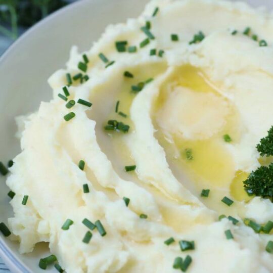 A bowl of fluffy white mashed potatoes topped with melting butter, fresh green chives, and parsley garnish.