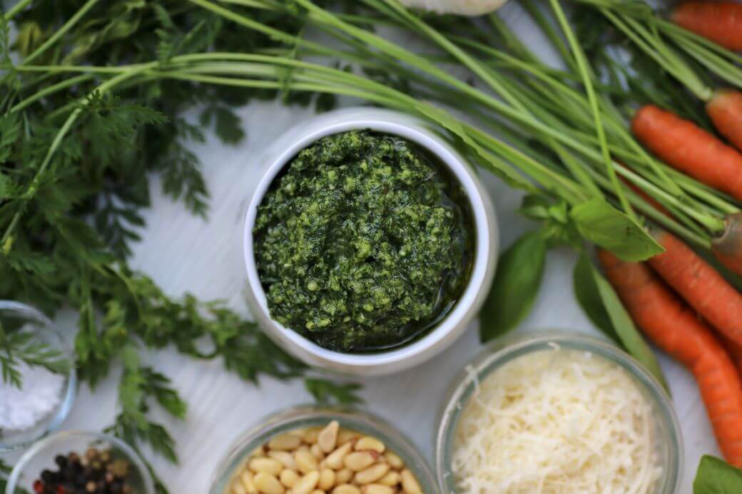 Green pesto in white bowl surrounded by whole carrots and ingredients.