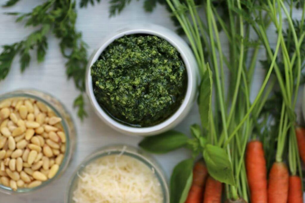 Green pesto in white bowl surrounded by whole carrots and ingredients.