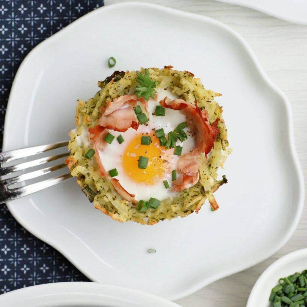 A baked yellow yolked egg with ham sits in a nest of hash browns.