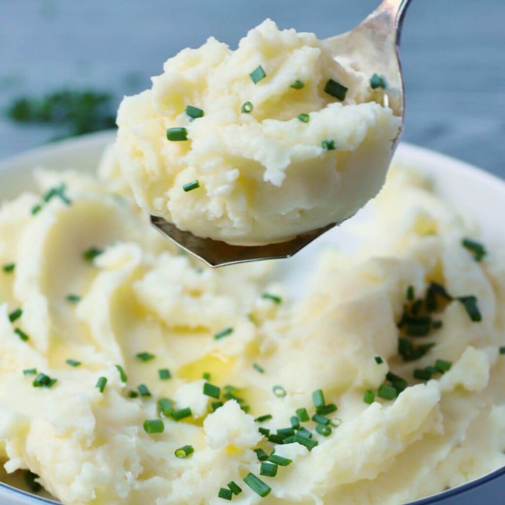 A silver spoon holds a serving of fluffy mashed potatoes over a bowl.