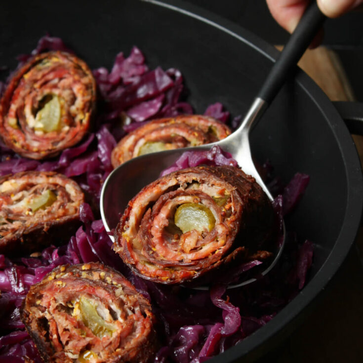 One Beef and bacon roll in a spoon above a pan of more meat rolls sitting on a bed of red cabbage.