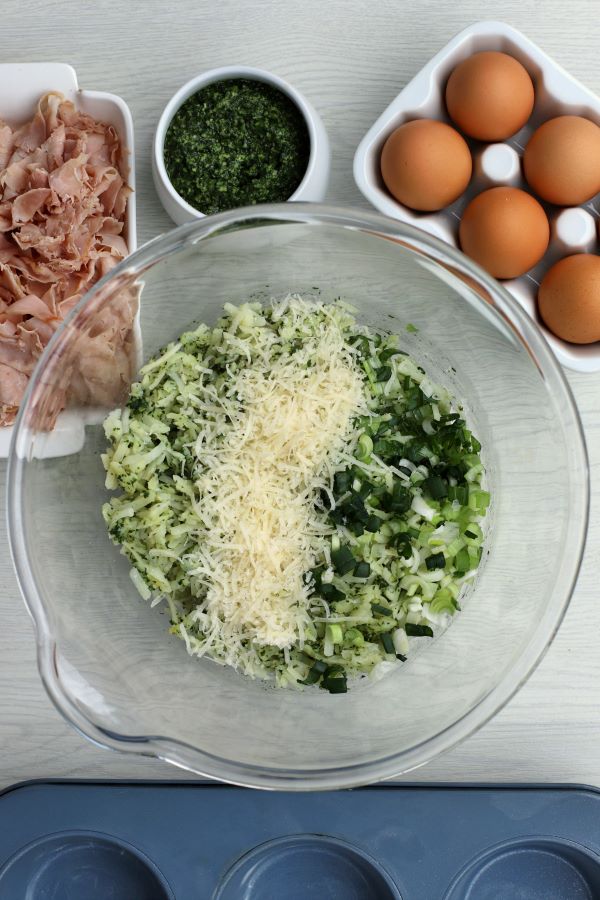 A glass bowl containing hash browns, pesto, cheese, and green onions surrounded by a tray of brown eggs, a bowl of pesto, and ham.