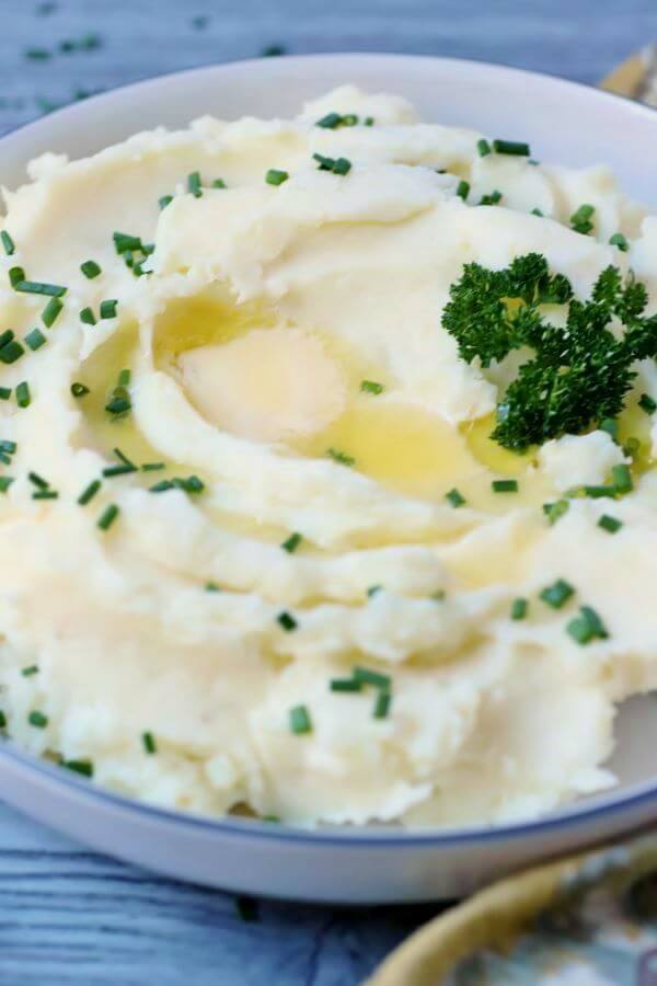 A bowl of fluffy white mashed potatoes topped with melting butter, fresh green chives, and parsley garnish.