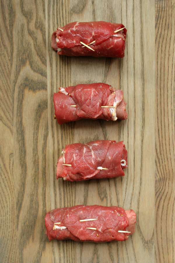 Four raw beef rolls on a wooden cutting board.