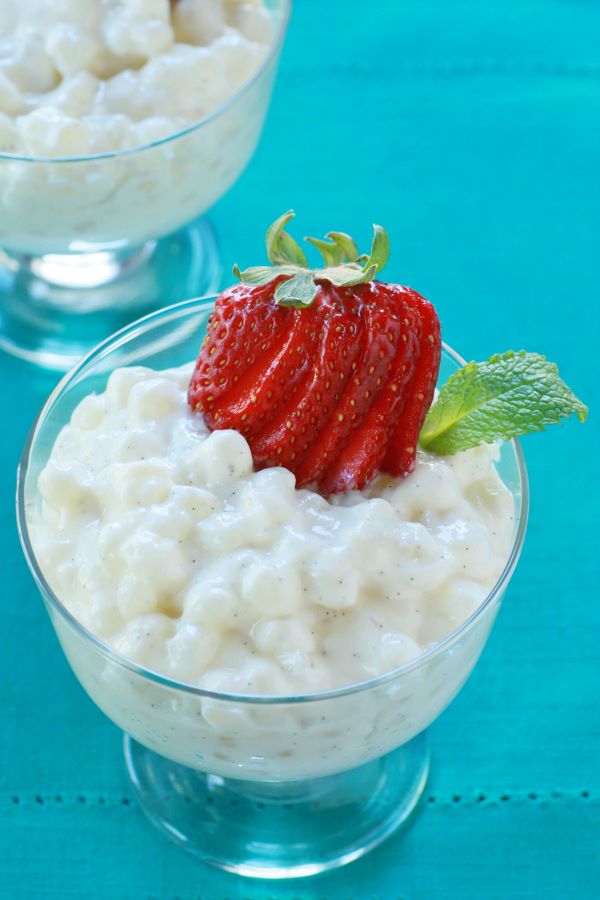 A bowl of creamy tapioca pudding topped with sliced strawberries on a aqua background.