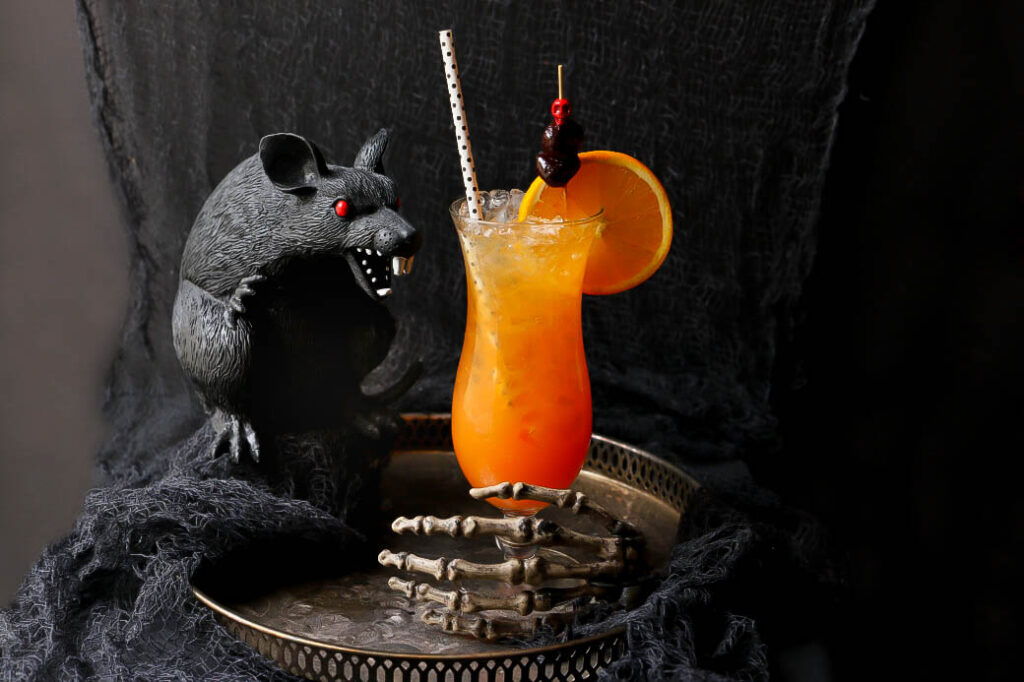 A vibrant orange cocktail garnished with a slice of orange and cherries on a striped straw, sits on a tray held by a skeleton hand. A menacing black rat statue with red eyes stands next to the drink against a dark, textured backdrop.