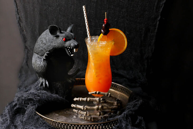A dark scene featuring a plastic black rat and an icy bright orange cocktail in a tall glass.