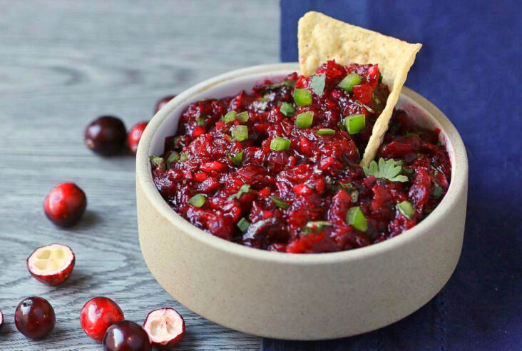 A tortilla chip in a white bowl filled with bright red cranberry salsa topped with fresh chopped cilantro and jalapenos.