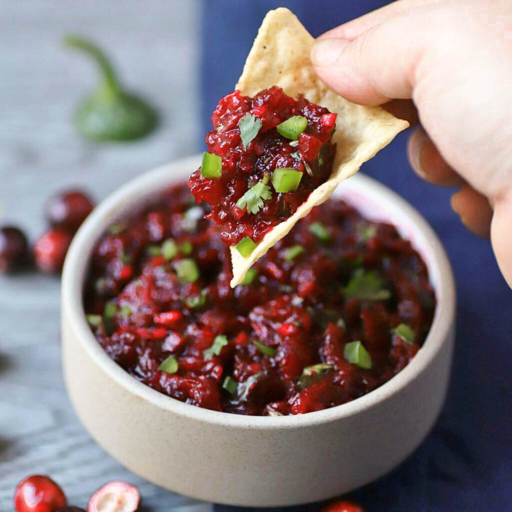 A hand dips a tortilla chip into a white bowl filled with bright red cranberry salsa topped with fresh chopped cilantro and jalapenos.