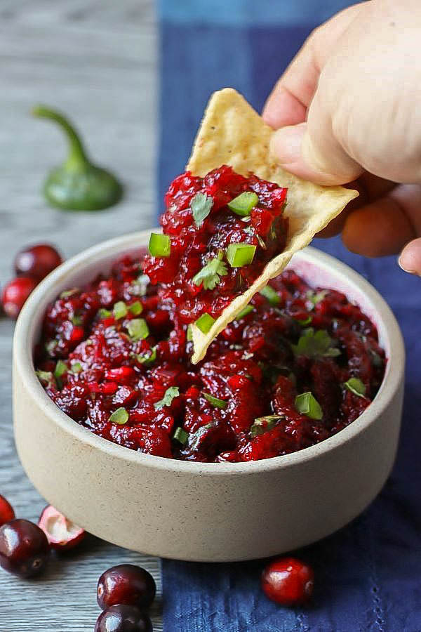 A hand dips a tortilla chip into a white bowl filled with bright red cranberry salsa topped with fresh chopped cilantro and jalapenos.