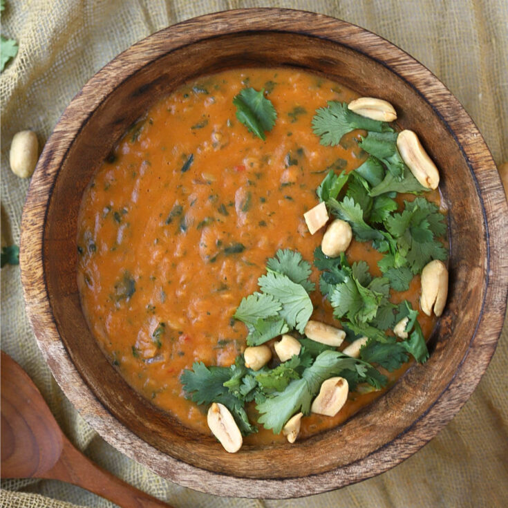 A creamy orange soup in a wooden bowl flecked with greens and garnished with cilantro and peanuts.