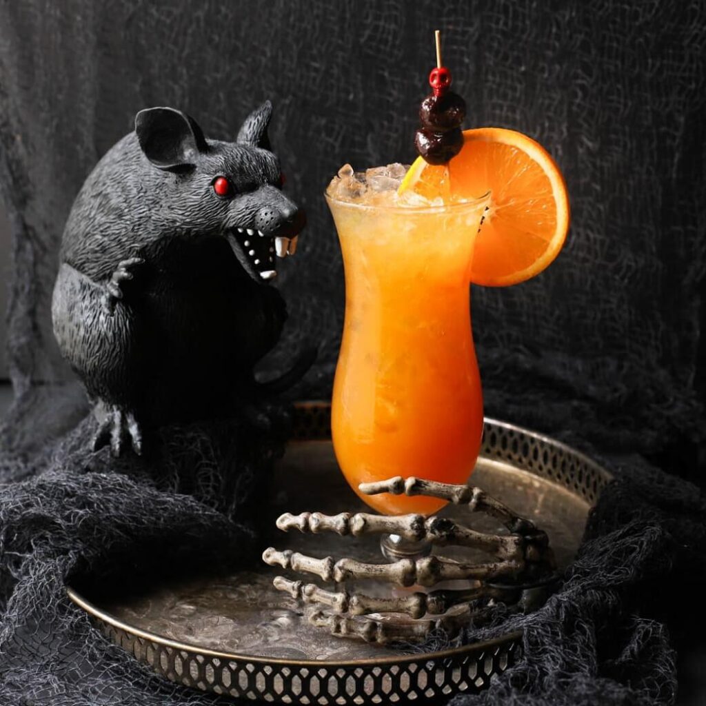 A dark scene featuring a plastic black rat and an icy bright orange cocktail in a tall glass.