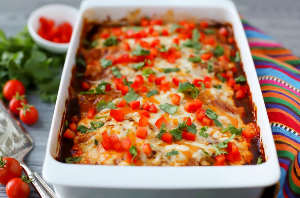 A white baking dish full of cheesy baked enchiladas topped with cilantro and red peppers.
