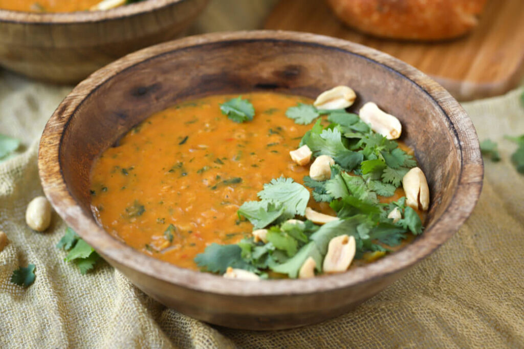A creamy orange soup in a wooden bowl flecked with greens and garnished with cilantro and peanuts.