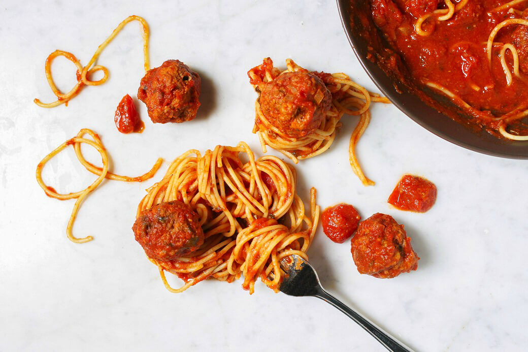 A group of saucy meatballs and coiled spaghetti on a white marble background.