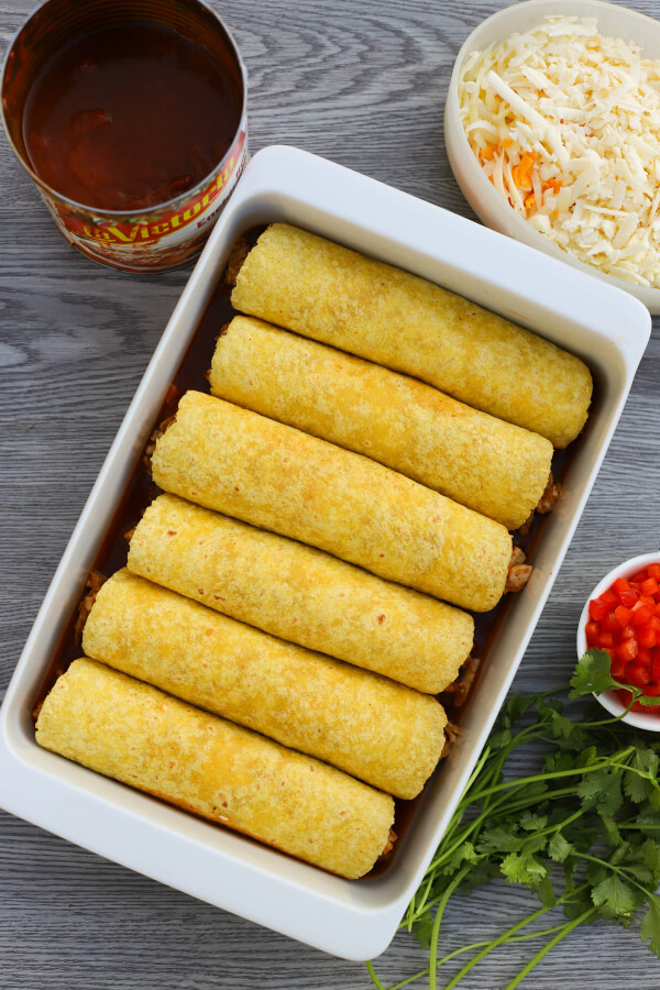 A white baking dish filled with rolled corn tortillas surrounded by sauce, cheese, cilantro, and diced red peppers.