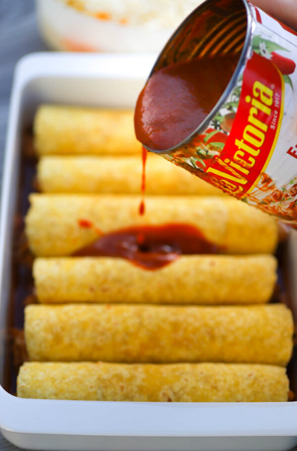Red enchilada sauce being drizzled onto a baking dish filled with rolled corn tortillas.
