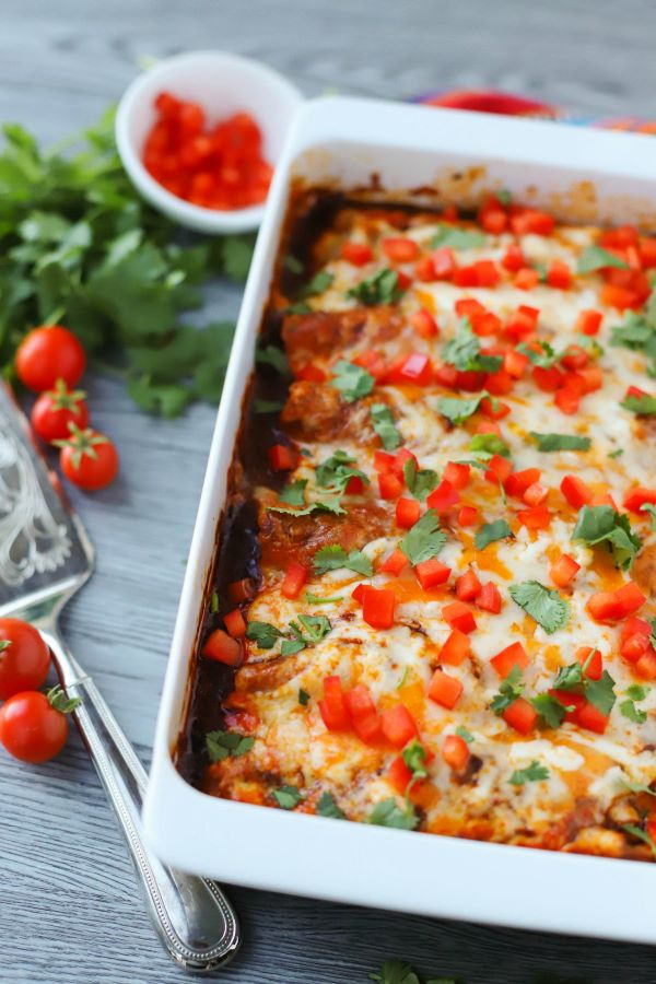 A white baking dish full of cheesy baked enchiladas topped with cilantro and red peppers.