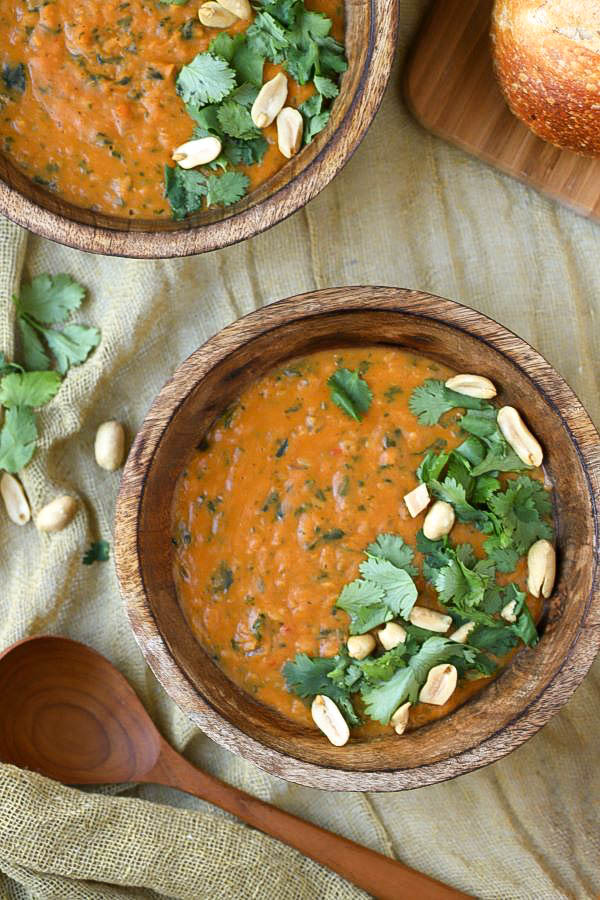 Two bowls of creamy orange vegan pumpkin soup in a wooden bowl flecked with greens and garnished with cilantro and peanuts.