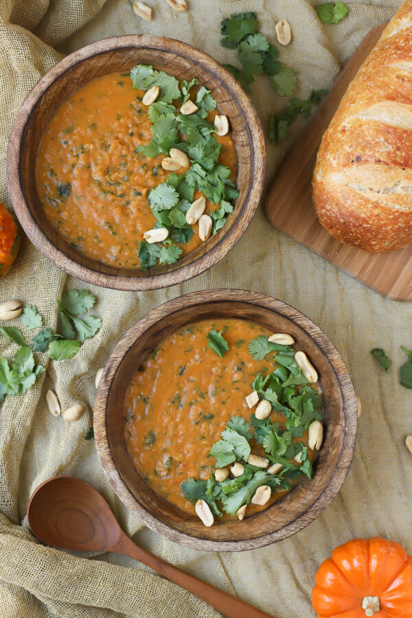 Two bowls of creamy orange vegan pumpkin soup in a wooden bowl flecked with greens and garnished with cilantro and peanuts.