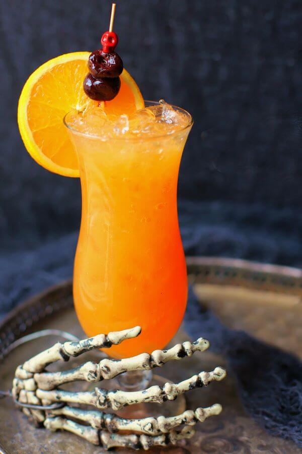 A dark scene featuring an icy bright orange cocktail in a tall glass held by a skeleton hand.