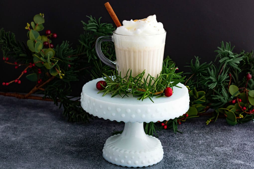 Un mug en verre rempli de lait de poule bourbon crémeux avec un bâton de cannelle sur un piédestal blanc.