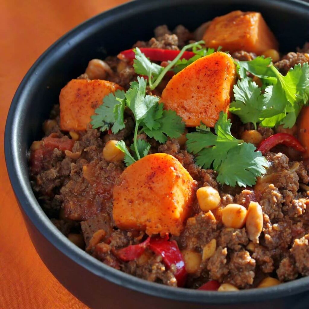 A black bowl filled with ground bison chili, peppers, chick peas, and large orange sweet potato bites.