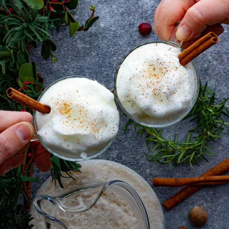 Two hands click glasses filled with bourbon eggnog together in holiday cheers.