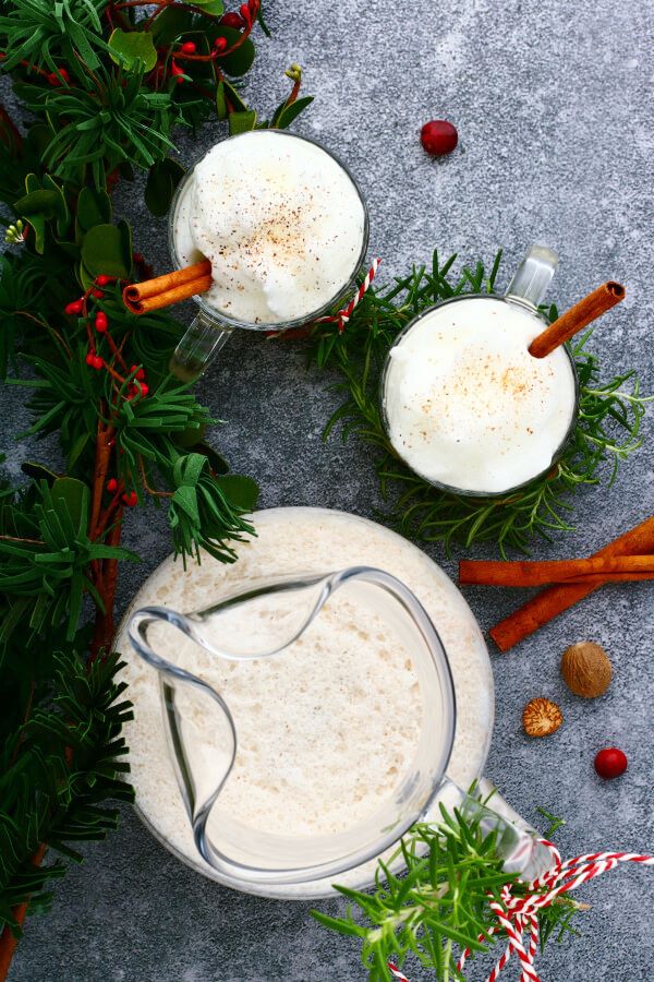 Two glass mugs and a pitcher filled with creamy eggnog topped with foam and grated nutmeg.