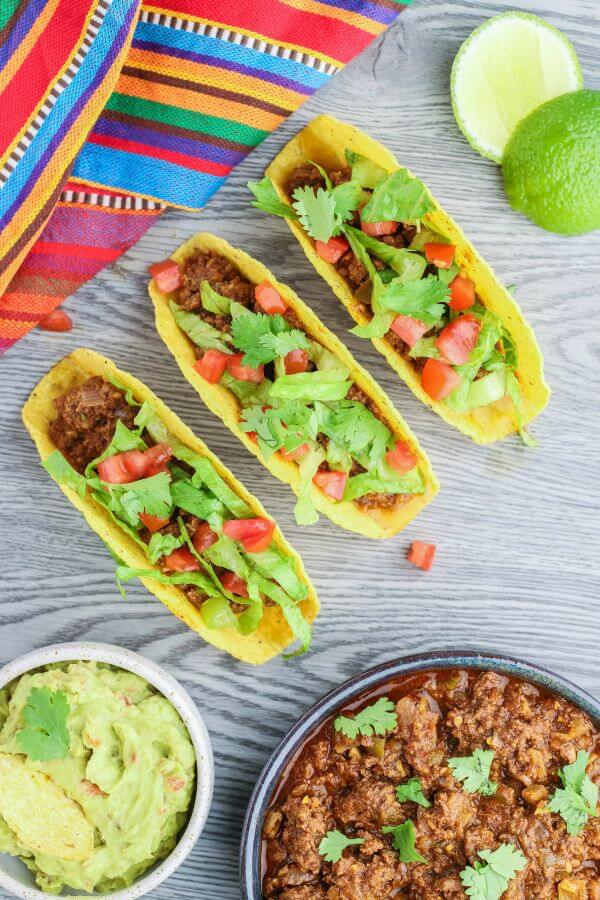 Three filled hard shell tacos surrounded by lime wedges, guacamole and a bowl of crockpot taco meat.