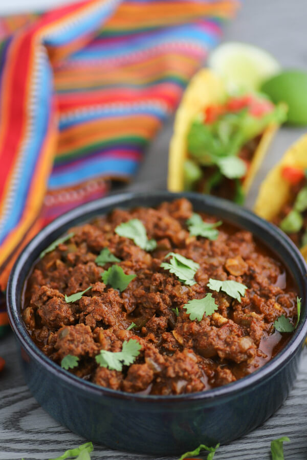 A bowl full of cooked spiced ground beef beside tacos and guacamole.