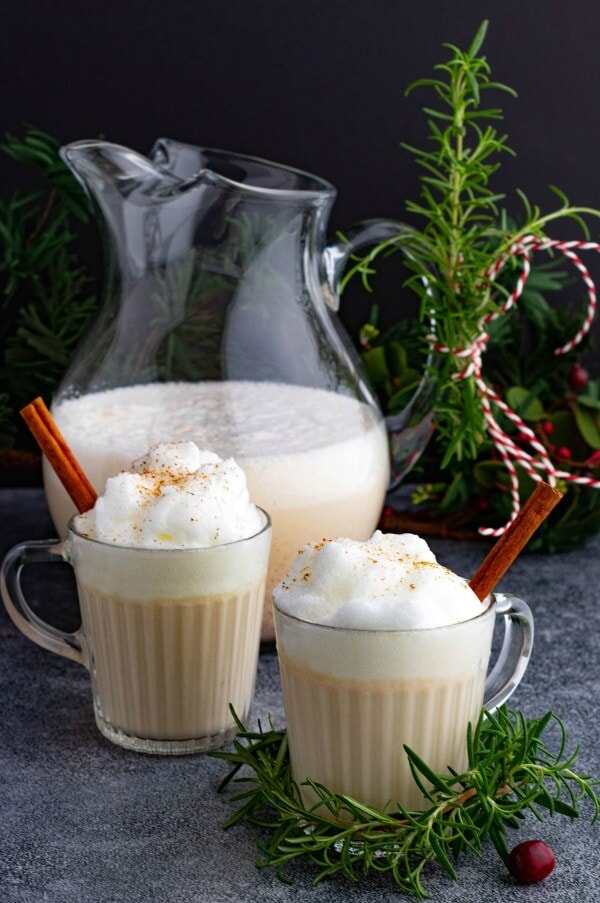 Two glass mugs and a pitcher filled with creamy eggnog topped with foam and grated nutmeg. 