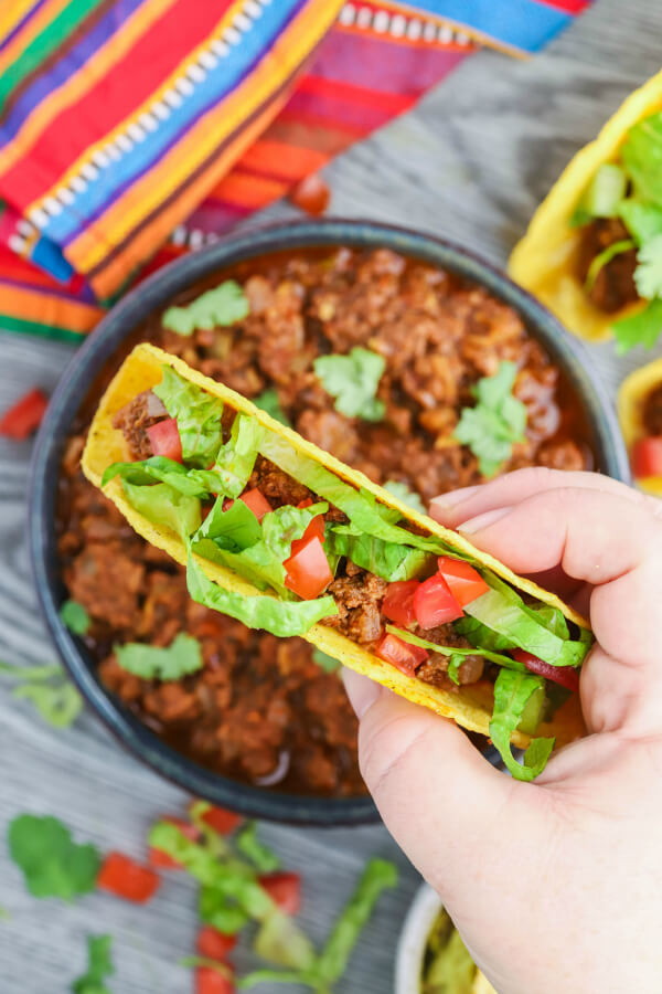 a hand holding a hard shell stuffed taco over a bowl of cooked crockpot taco meat.