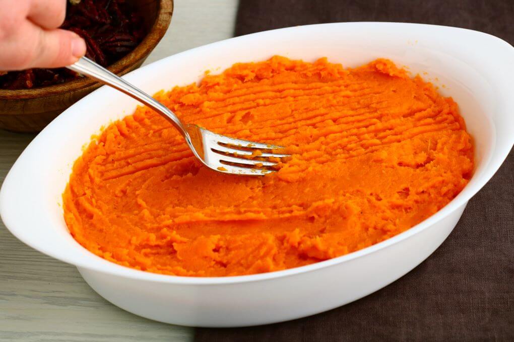 A fork lays down a design on a bowl of bright orange mashed sweet potatoes.