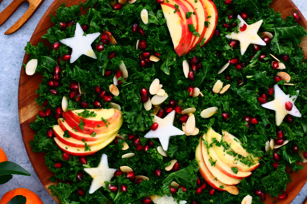A leafy green massaged kale salad topped with apple slices, daikon stars, almonds, and pomegranate seeds.