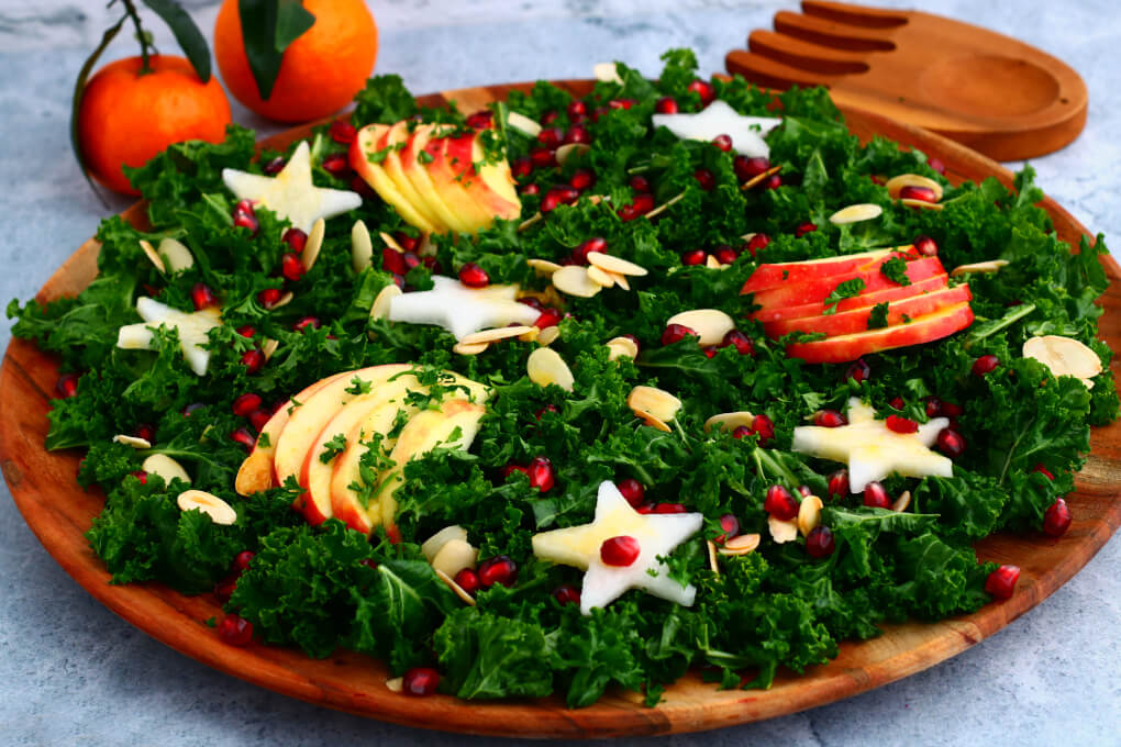 A leafy green massaged kale salad topped with apple slices, daikon stars, almonds, and pomegranate seeds.