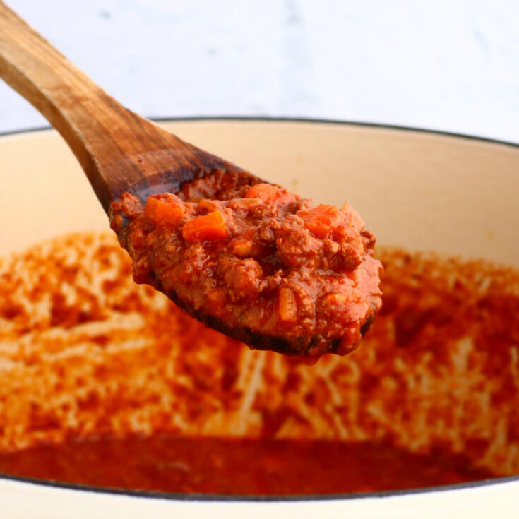 A wooden spoon holds bright red rich meat sauce over a pot of sauce.