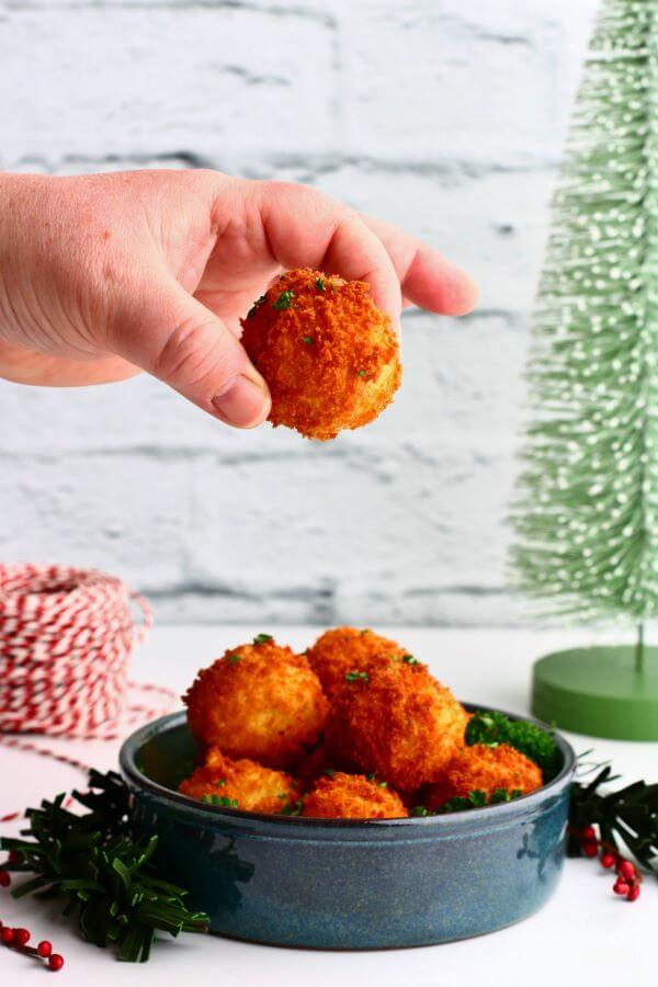 A hand holding a golden fried goat cheese ball above a bowl full of fried cheese balls.