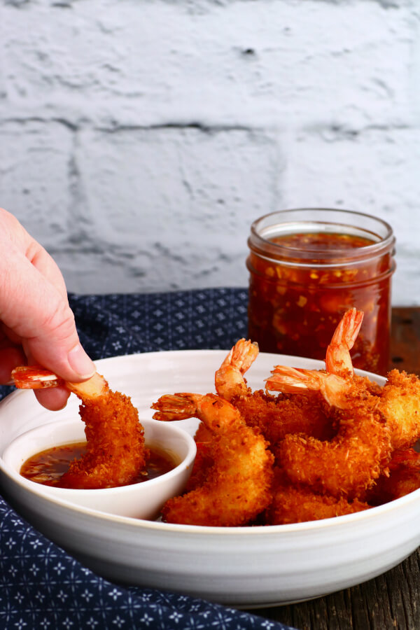 A hand dips a golden fried shrimp into a bowl of red chili sauce.