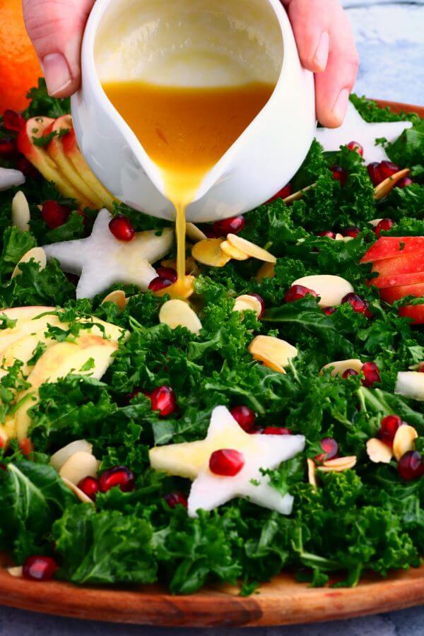 Mandarin orange vinaigrette being poured over a leafy green massaged kale salad topped with apple slices, daikon stars, almonds, and pomegranate seeds.